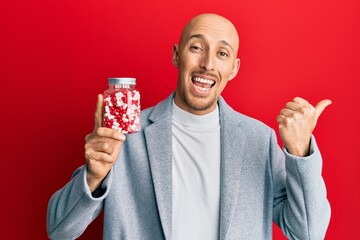Bald man with beard holding jar full of pills pointing thumb up to the side smiling happy with open mouth
