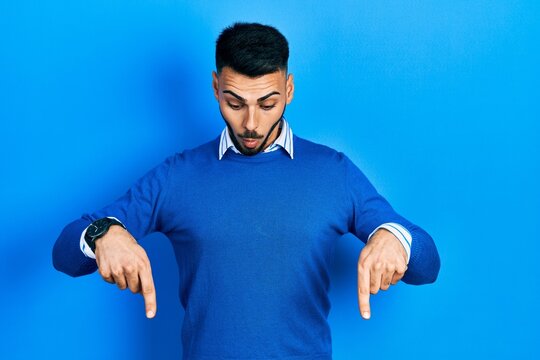 Young hispanic man with beard wearing casual blue sweater pointing down with fingers showing advertisement, surprised face and open mouth