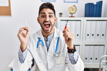 Young doctor man holding electronic cigarette at medical clinic celebrating crazy and amazed for success with open eyes screaming excited.
