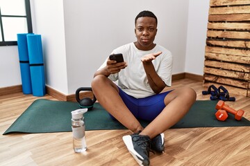Obraz premium Young african man sitting on training mat at the gym using smartphone looking at the camera blowing a kiss with hand on air being lovely and sexy. love expression.