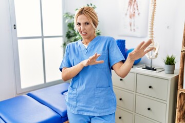 Beautiful blonde physiotherapist woman working at pain recovery clinic disgusted expression, displeased and fearful doing disgust face because aversion reaction. with hands raised