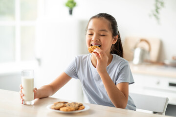 Pretty asian girl eating cookies and drinking milk in kitchen, happy female child enjoying healthy snack at home