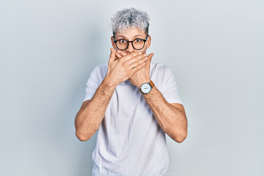 Young hispanic man with modern dyed hair wearing white t shirt and glasses shocked covering mouth with hands for mistake. secret concept.