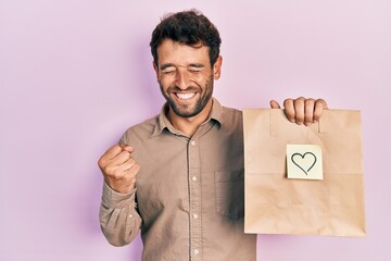 Handsome man with beard holding delivery paper bag with heart reminder very happy and excited doing...