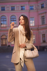 Fototapeta premium Young beautiful elegant woman outdoor portrait. Fashionable girl in beige blazer, trousers, white shoes and scarf. Lady with long brunette straight hair walking at city street