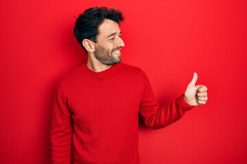Handsome man with beard wearing casual red sweater looking proud, smiling doing thumbs up gesture to the side