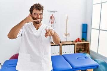 Young handsome physiotherapist man working at pain recovery clinic smiling doing talking on the telephone gesture and pointing to you. call me.