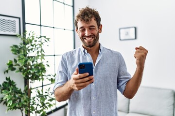 Young handsome man using smartphone at home screaming proud, celebrating victory and success very...