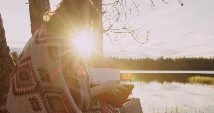 Young girl with red golden hair flips through a sketchbook during sunset in the forest, sits on the shore of a lake. Sunset sunlight on ginger hair