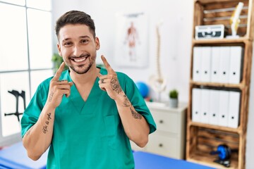 Young physiotherapist man working at pain recovery clinic smiling with open mouth, fingers pointing...