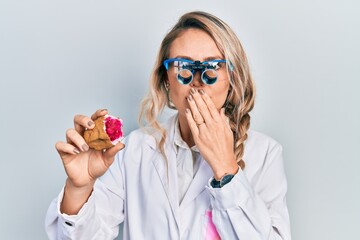 Beautiful young blonde woman holding geode stone wearing magnifier glasses covering mouth with hand, shocked and afraid for mistake. surprised expression