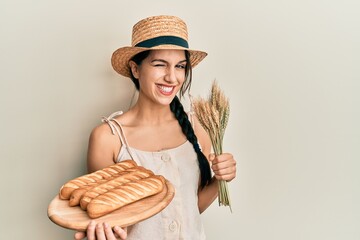 Young hispanic woman holding homemade bread and spike wheat winking looking at the camera with sexy...