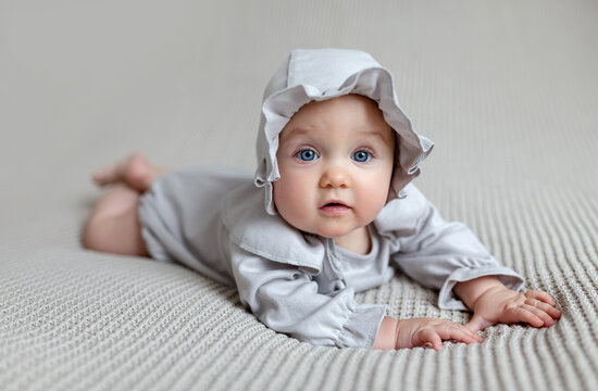 Happy cute baby in a white light bodysuit and hat on the blanket at home looking at camera