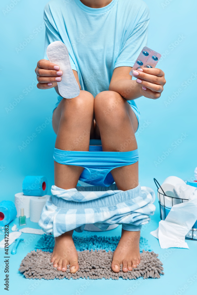 Wall mural Unrecognizable woman sits on wc bowl has menses holds sanitary napkin and pills to reveal pain suffers from period cramps keeps feet on carpet poses in washroom. Daily necessities. Female hygiene