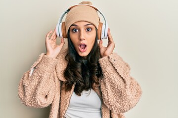 Young hispanic woman listening to music using headphones afraid and shocked with surprise and amazed expression, fear and excited face.