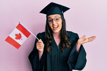 Young hispanic woman wearing graduation uniform holding canada flag celebrating achievement with happy smile and winner expression with raised hand