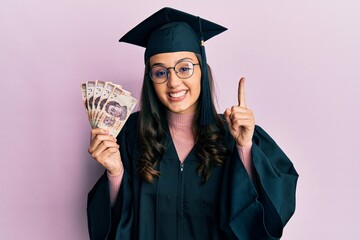 Young hispanic woman wearing graduation uniform holding mexican pesos banknotes smiling with an...