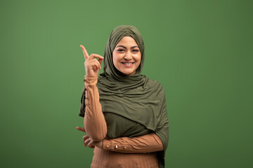 Positive islamic lady in hijab having idea, pointing finger up, having inspirational moment over olive background