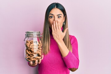 Young hispanic woman holding jar of chocolate chips cookies covering mouth with hand, shocked and afraid for mistake. surprised expression