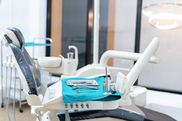 Side view selective focus of dental instruments set placed on a medical disposable blue napkin on an operator's table near an empty dental chair with a blurred bright dental clinic room in background.