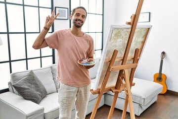 Young hispanic man with beard painting on canvas at home smiling looking to the camera showing fingers doing victory sign. number two.