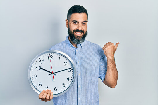 Young hispanic man holding big clock pointing thumb up to the side smiling happy with open mouth
