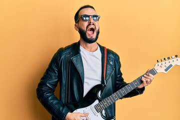 Young man with beard playing electric guitar angry and mad screaming frustrated and furious,...