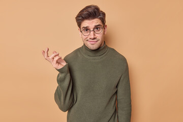 Puzzled confused man with dark hair has doubtful expression makes choice shrugs shoulders hesitates...