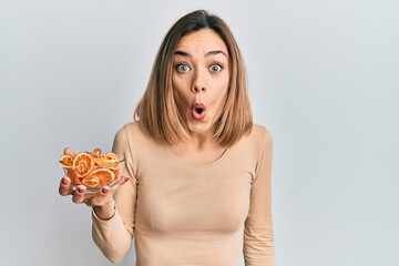 Young caucasian blonde woman holding bowl of dry orange scared and amazed with open mouth for surprise, disbelief face