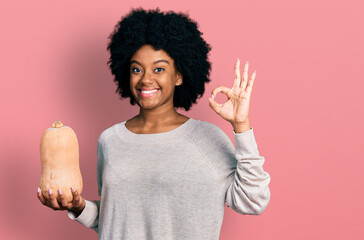 Young african american woman holding healthy fresh pumpkin doing ok sign with fingers, smiling friendly gesturing excellent symbol