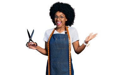 Young african american woman dressmaker designer wearing atelier apron holding scissors celebrating...