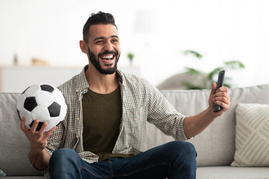 Happy young Arab man watching soccer game on television, holding ball and controller at home