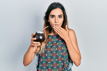 Young hispanic girl drinking mate infusion covering mouth with hand, shocked and afraid for...
