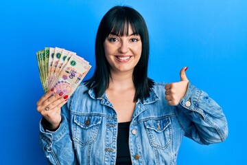 Young hispanic woman holding argentine pesos banknotes smiling happy and positive, thumb up doing...