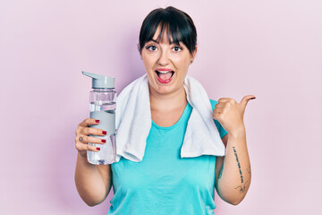 Young hispanic woman wearing sportswear holding water bottle pointing thumb up to the side smiling happy with open mouth