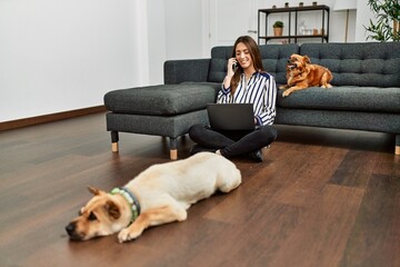 Young hispanic woman using laptop and talking on the smartphone sitting on floor with dogs at home