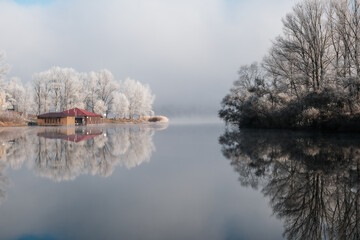 House by the lake. The transition of nature from autumn to winter. Foggy and sunny morning on the lake. A rare natural phenomenon. Fog over the lake and ripples on trees and grass.