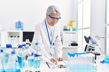 Middle age grey-haired woman wearing scientist uniform writing on clipboard working at laboratory