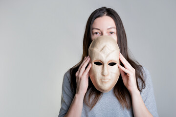 A young woman holds a mask in her hands, hiding her face half, a sly look over the mask. Photos in light gray tones, gray background