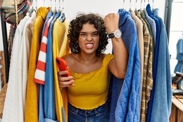 Young hispanic woman searching clothes on clothing rack using smartphone angry and mad raising fist...