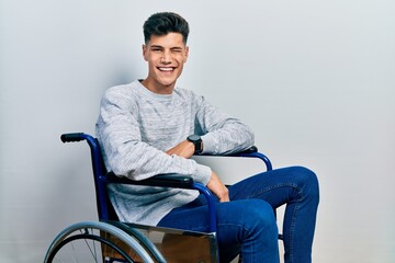 Young hispanic man sitting on wheelchair winking looking at the camera with sexy expression,...