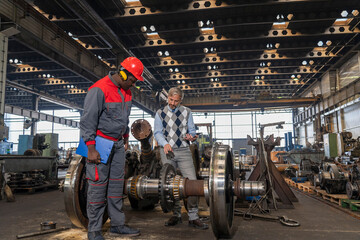 Fototapeta premium Production Manager Talking To African American Worker In Protective Workwear At Train Factory - Multiracial Industrial Co-Workers Standing Next To Train Wheels. Area For Train Maintenance And Repair