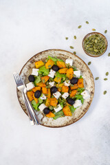 Healthy salad with fried pumpkin, feta cheese, olives, salad greens, pumpkin seeds on the light grey background.