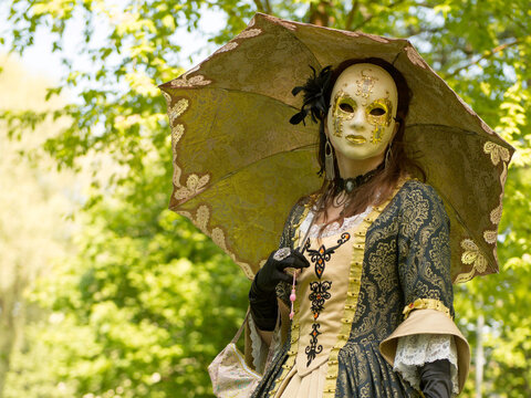 Costumed and masked woman at venetian carnival with golden umbrella. Blu sky and trees in the background.
