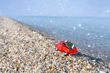 Red retro small car carrying christmas tree on the sea beach shore in snowfall. Winter holiday concept