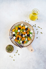 Healthy salad with fried pumpkin, feta cheese, olives, salad greens, pumpkin seeds, olive oil on the light grey background.