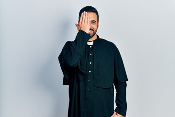 Handsome hispanic man with beard wearing catholic priest robe covering one eye with hand, confident...