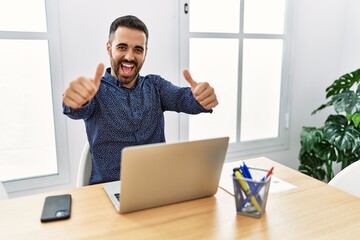 Young hispanic man with beard working at the office with laptop approving doing positive gesture with hand, thumbs up smiling and happy for success. winner gesture.