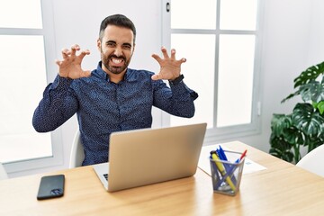 Young hispanic man with beard working at the office with laptop smiling funny doing claw gesture as...