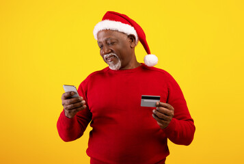 Cheerful senior black man with cellphone and credit card wearing Santa hat, buying Christmas gifts online